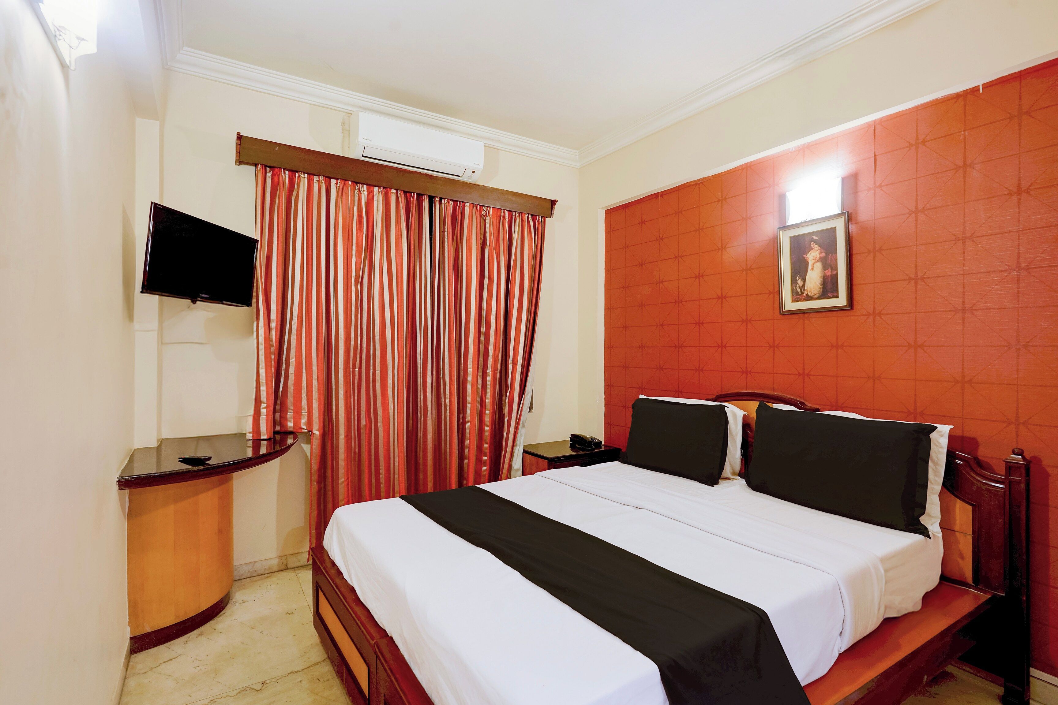 Habitación de hotel con cama, mesita de noche, televisor montado en la pared y ventana con cortinas.