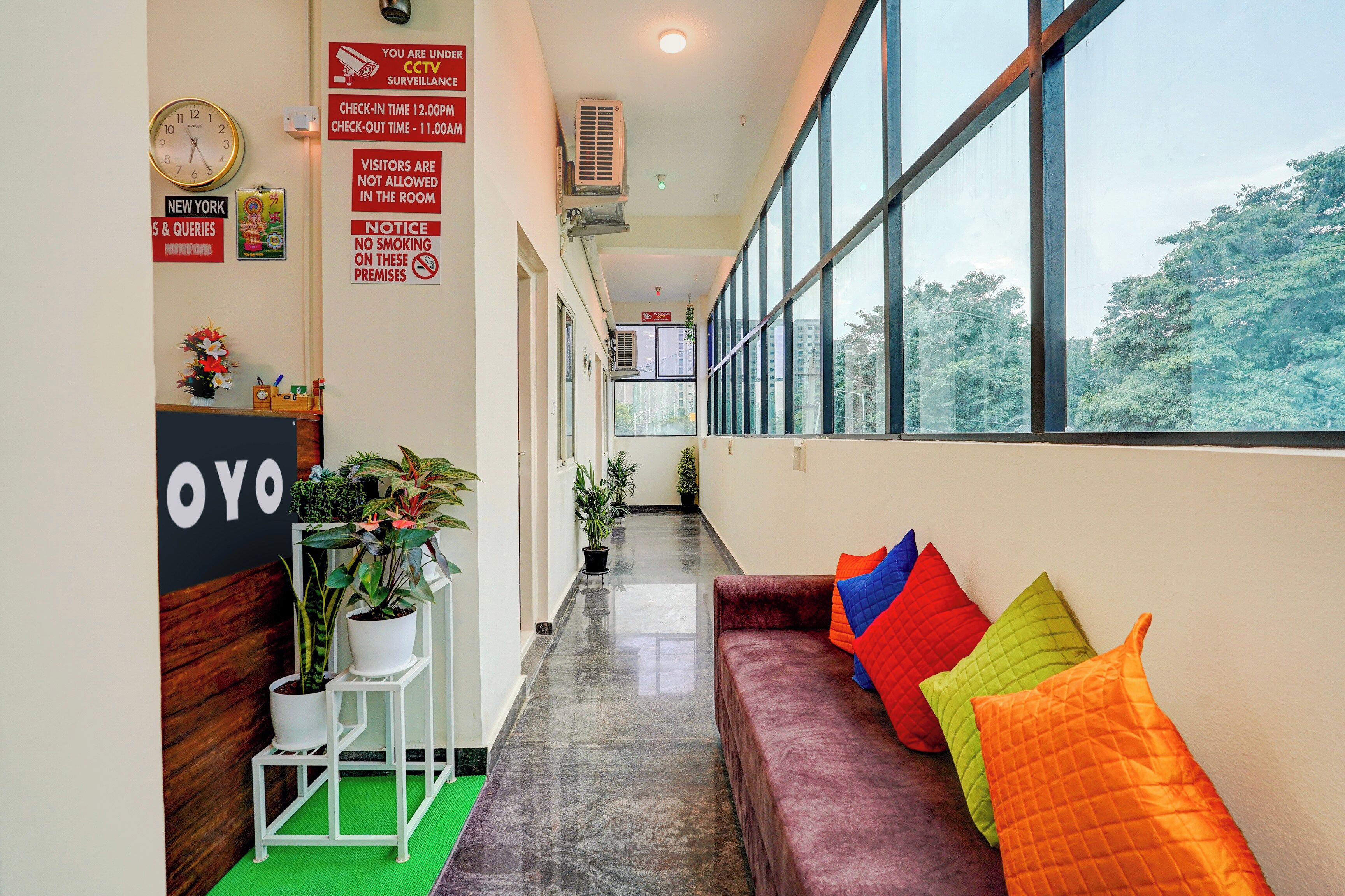 A hotel lobby with a reception desk, a clock, CCTV surveillance notice, and a sofa with colorful cushions.