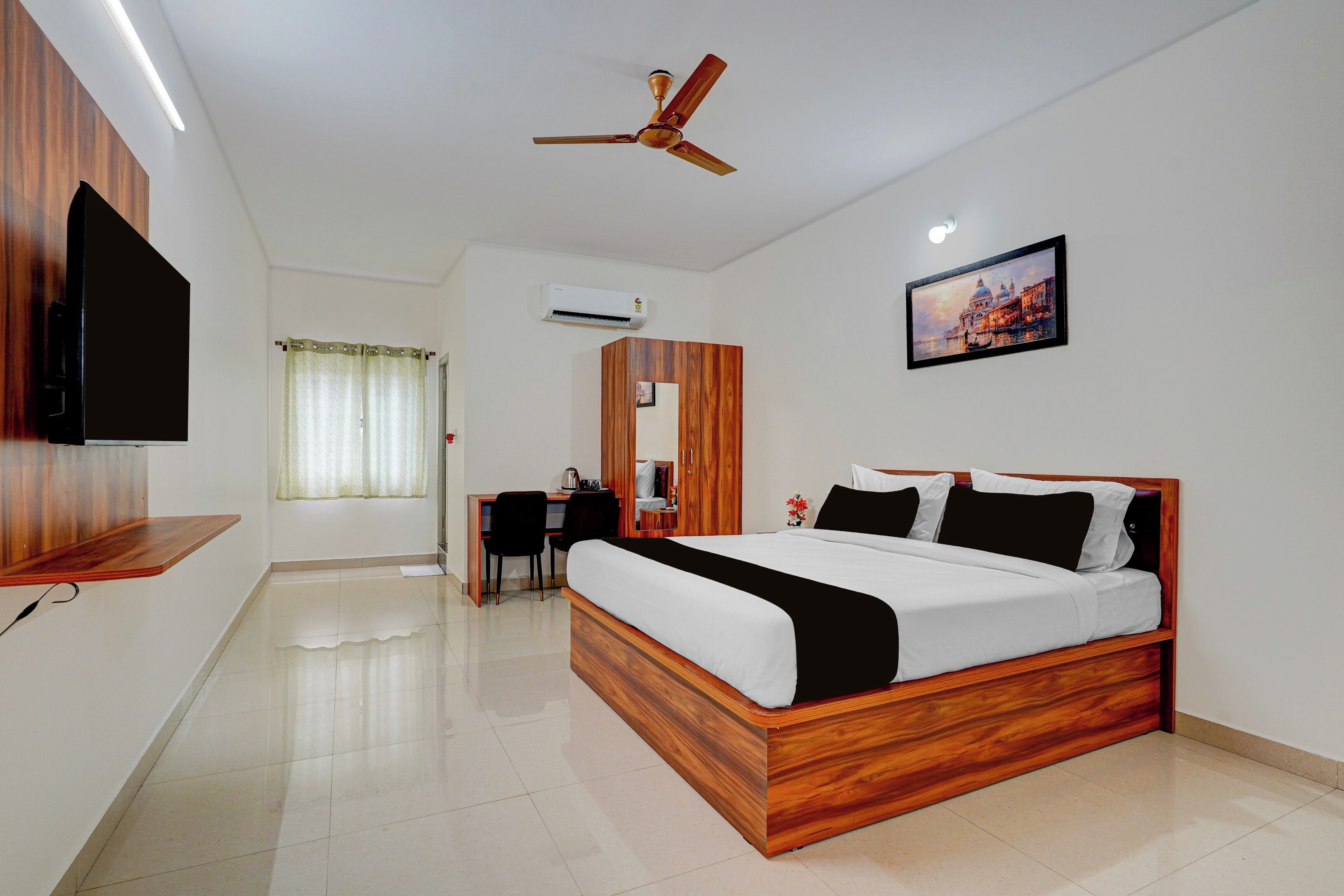 A hotel room with a wooden bed, a desk, a chair, a television, an air conditioning unit, and a painting on the wall.