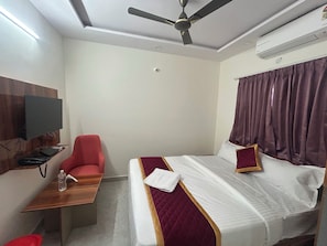 A hotel room with a bed, a desk with a chair, a television mounted on the wall, and a ceiling fan. - Hotel O Srinidhi Inn (Bangalore)