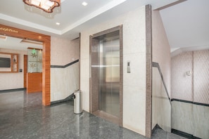 Un elevador moderno con detalles metálicos, un cesto de basura y un área de recepción con mostrador de madera y televisor de pared.