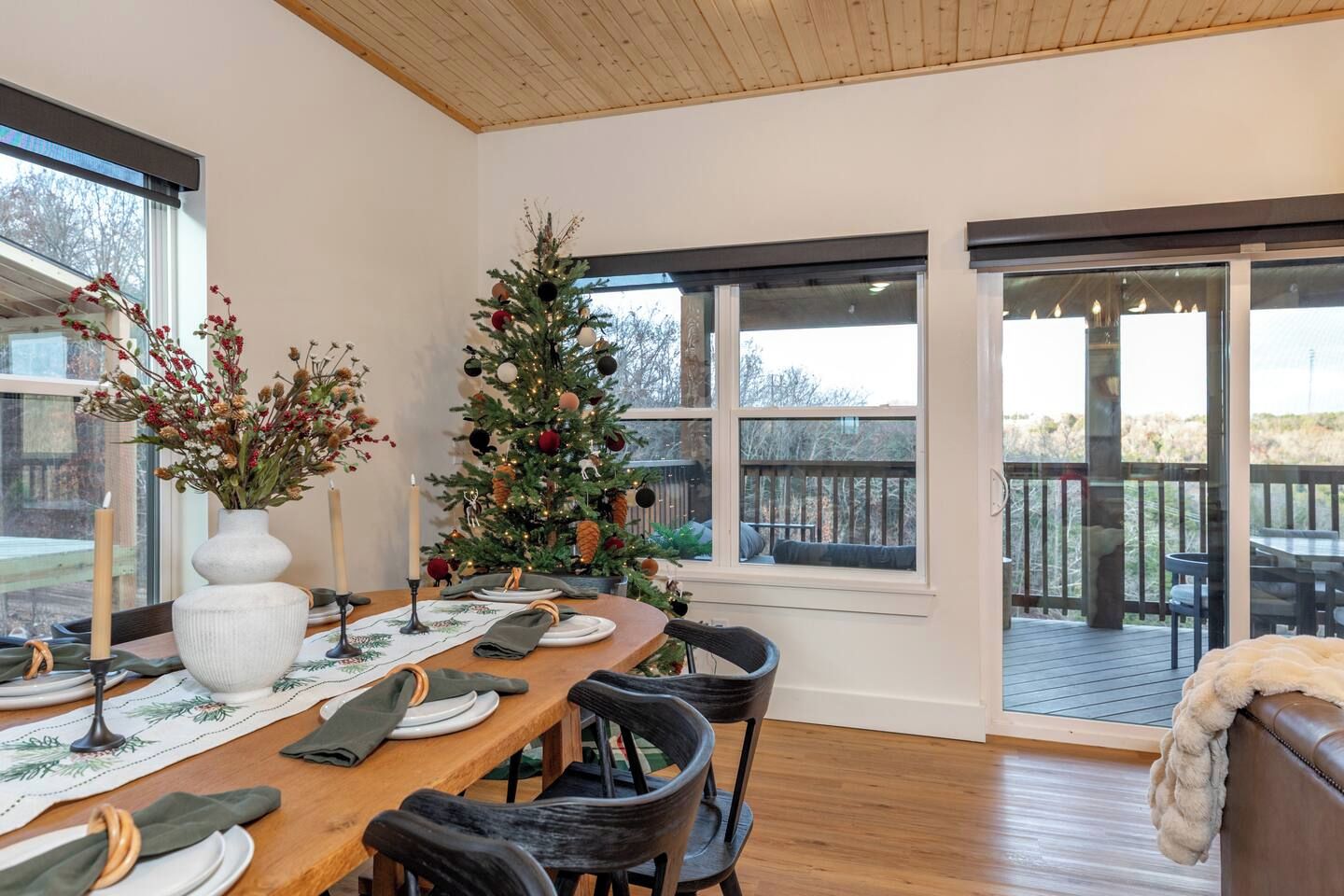 Un coin repas avec une table en bois dressée pour quatre personnes, un sapin de Noël et une vue sur l’extérieur grâce à de grandes fenêtres.