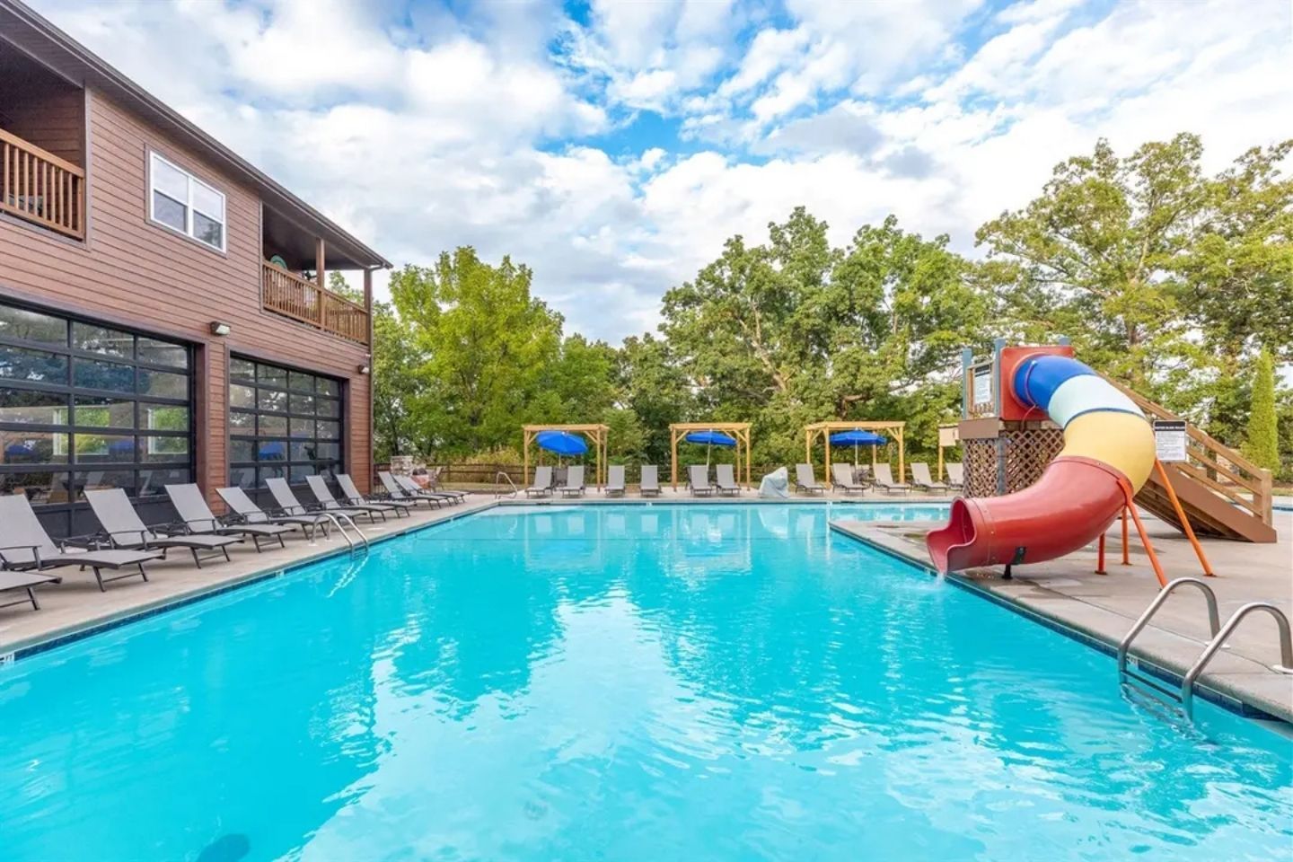 A swimming pool with lounge chairs and a slide, surrounded by trees and a building.