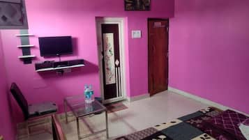 A pink-walled room with a flat-screen TV, a glass table, a sofa, and a patterned rug.