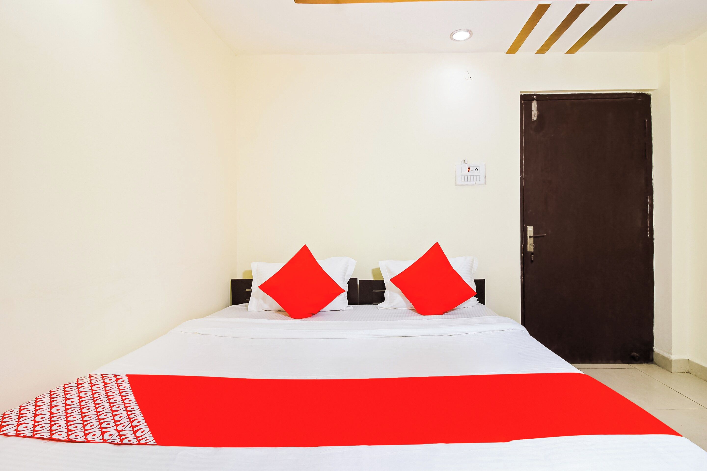 A hotel room with a bed, two red pillows, a white bedspread, a red and white patterned blanket, a wall socket, and a door.