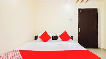 A hotel room with a bed, two red pillows, a white bedspread, a red and white patterned blanket, a wall socket, and a door.