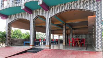 Un espacio de estar al aire libre con techo, con sillas y mesas rojas, rodeado por un muro con diseño y una escalera que sube a un balcón.