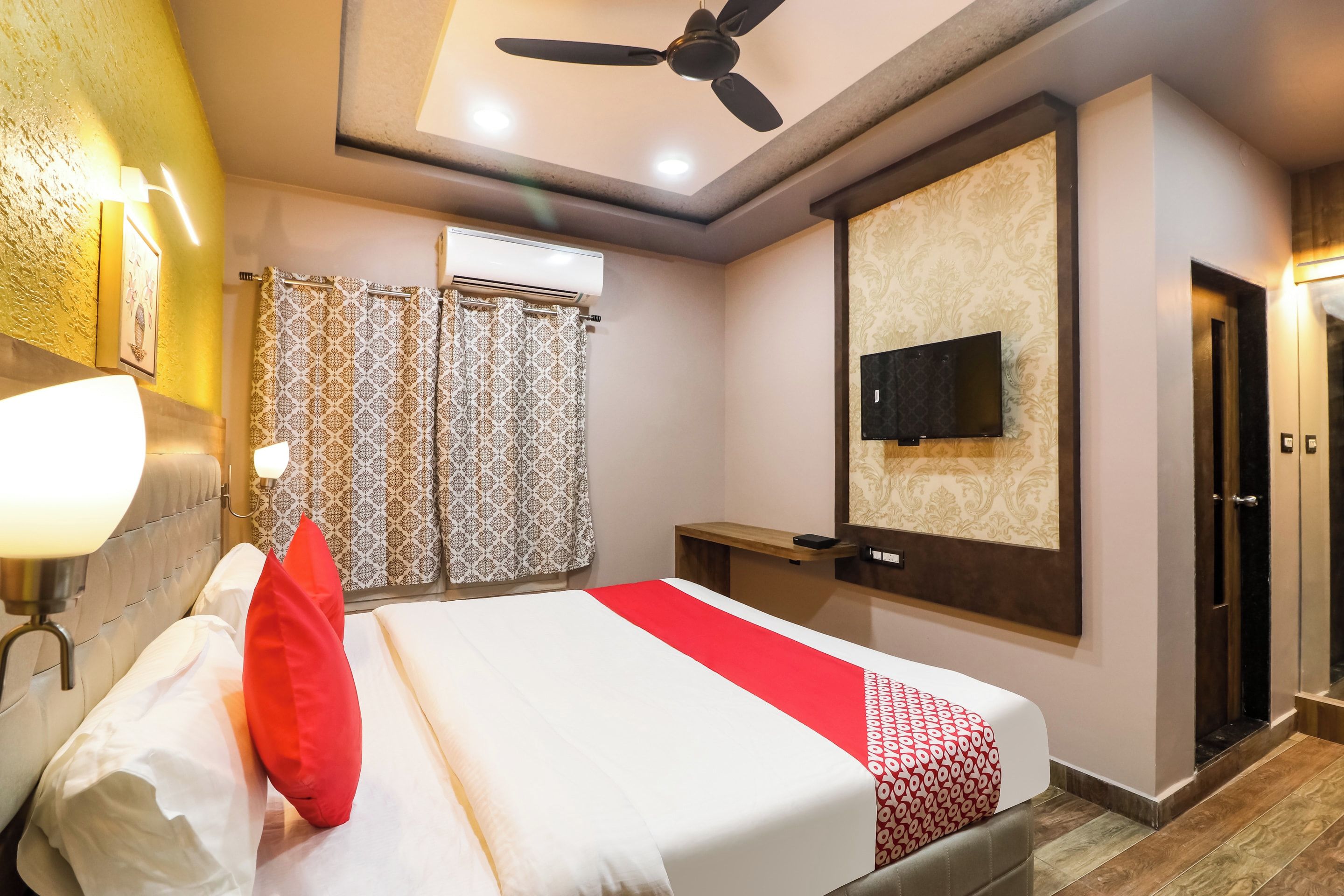 A hotel room with a bed, a ceiling fan, a TV mounted on the wall, and a shower area with patterned curtains.