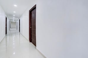 A long, narrow hallway with white walls and a shiny floor, leading to a door with a dark wooden frame. - Hotel O Viraj Luxury Stay (Warangal)