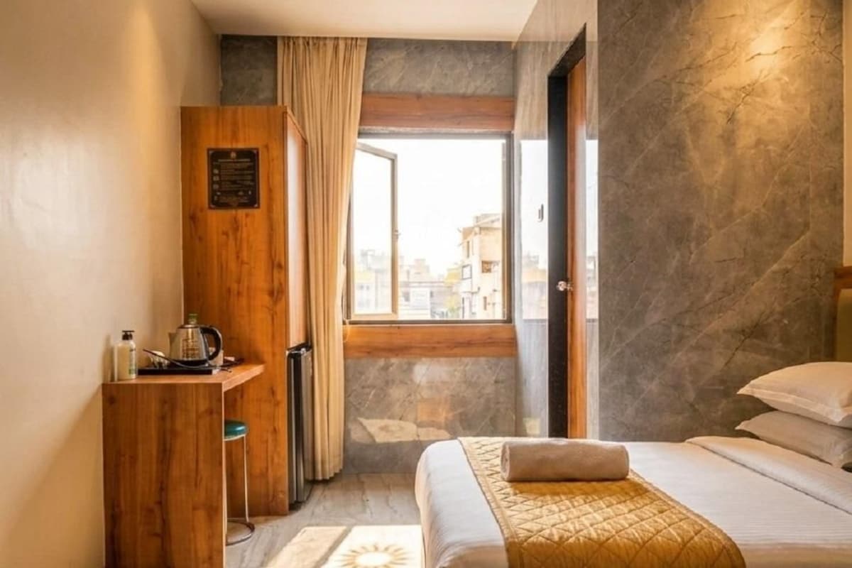 A hotel room with a bed, a wooden desk, a window with curtains, and a stone wall.