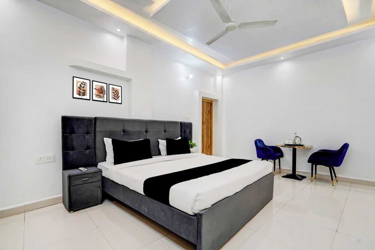 Quarto de hotel moderno com uma cama grande, mesas de cabeceira, ventoinha no teto e uma pequena área de estar com duas cadeiras e uma mesa.