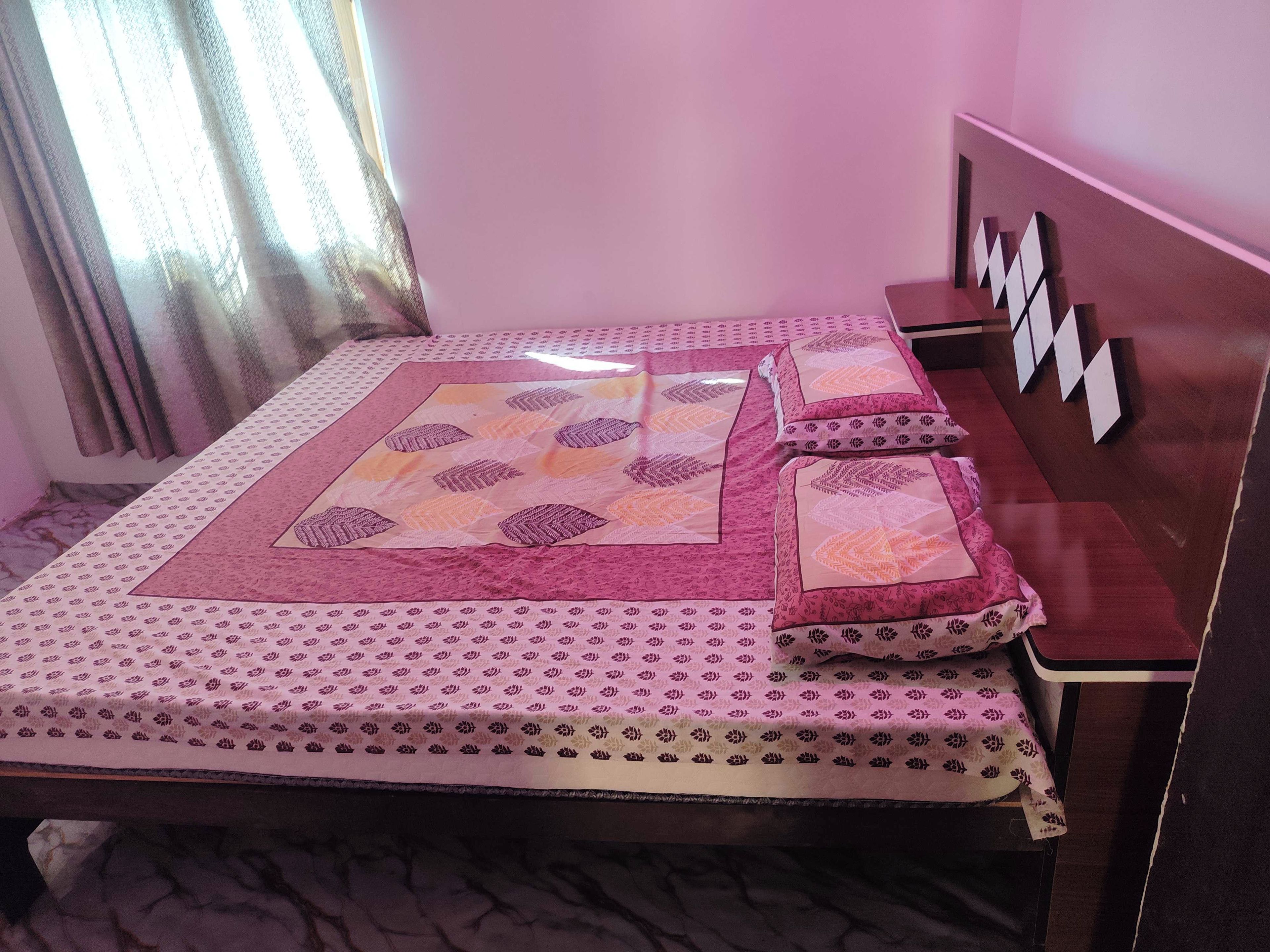 A bed with a patterned bedspread and pillows, a wooden headboard, and a window with curtains.