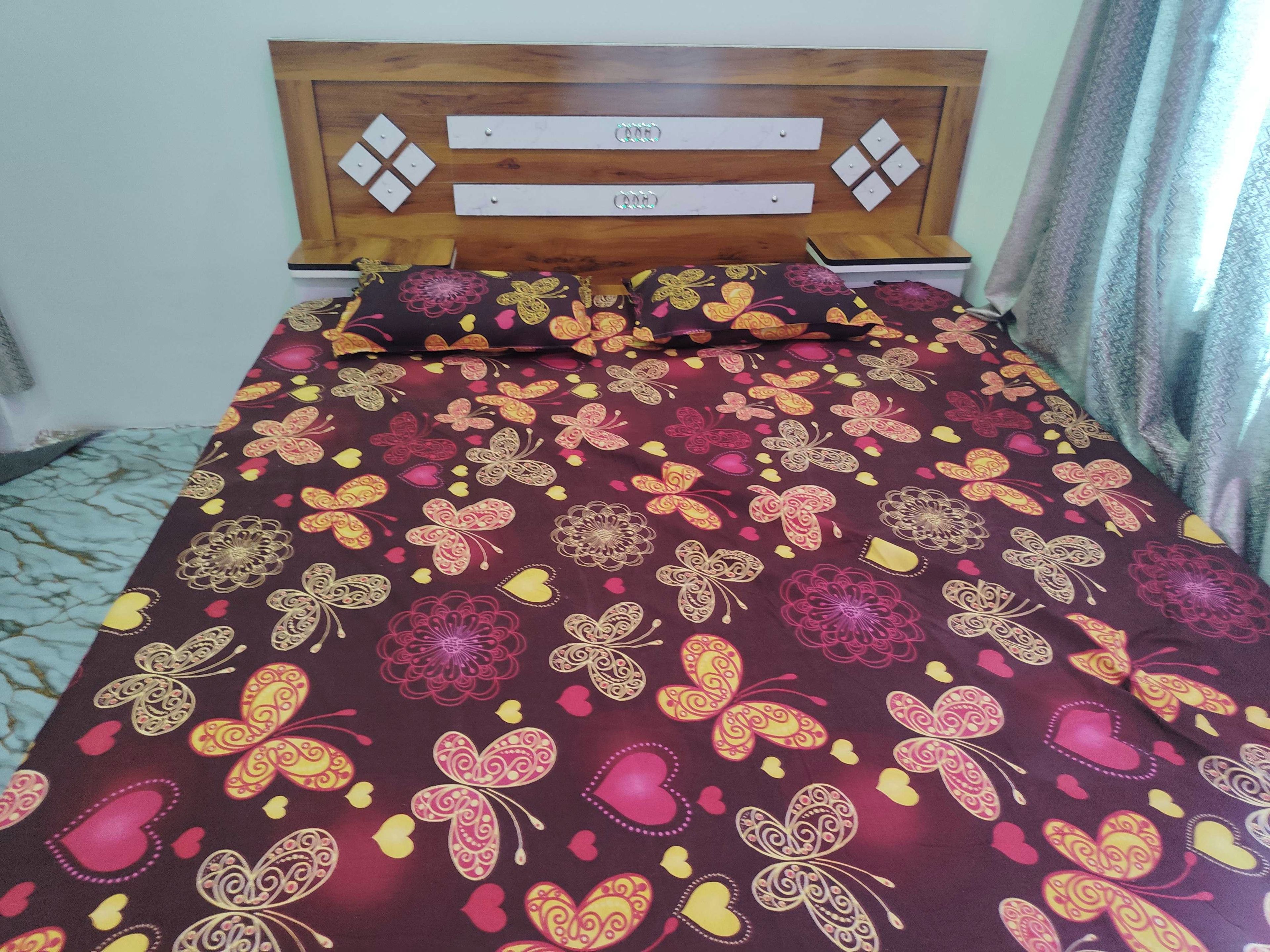 Una cama con cubrecama estampado con motivos florales y mariposas, cabecera de madera con controles de la luz y una ventana con cortinas estampadas.