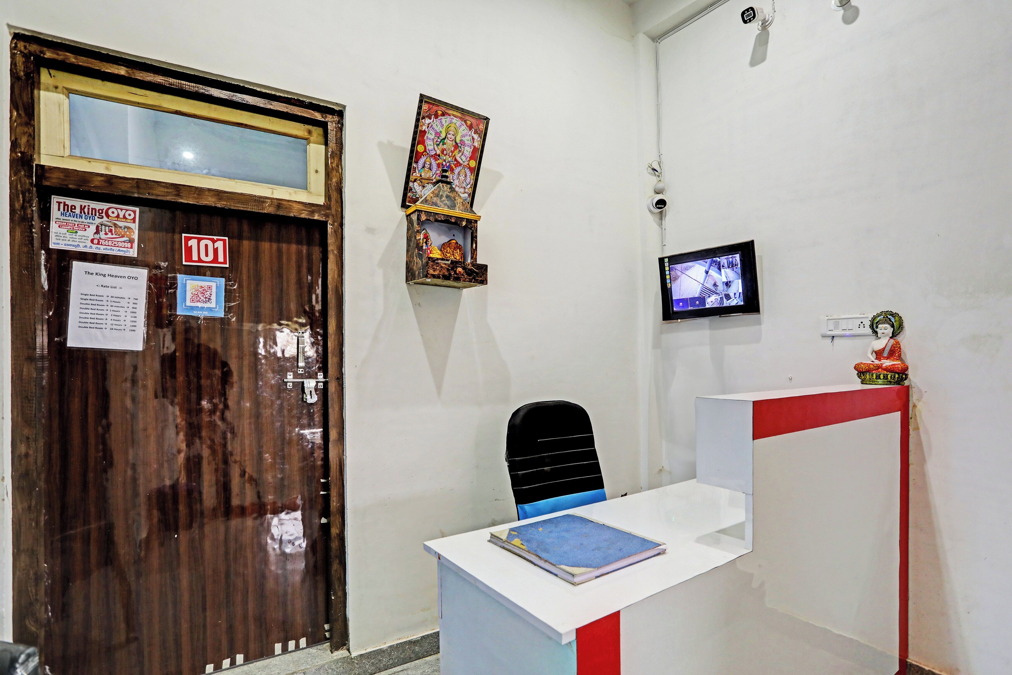 A hotel reception area with a wooden door, a reception desk, a CCTV camera, and a small statue.