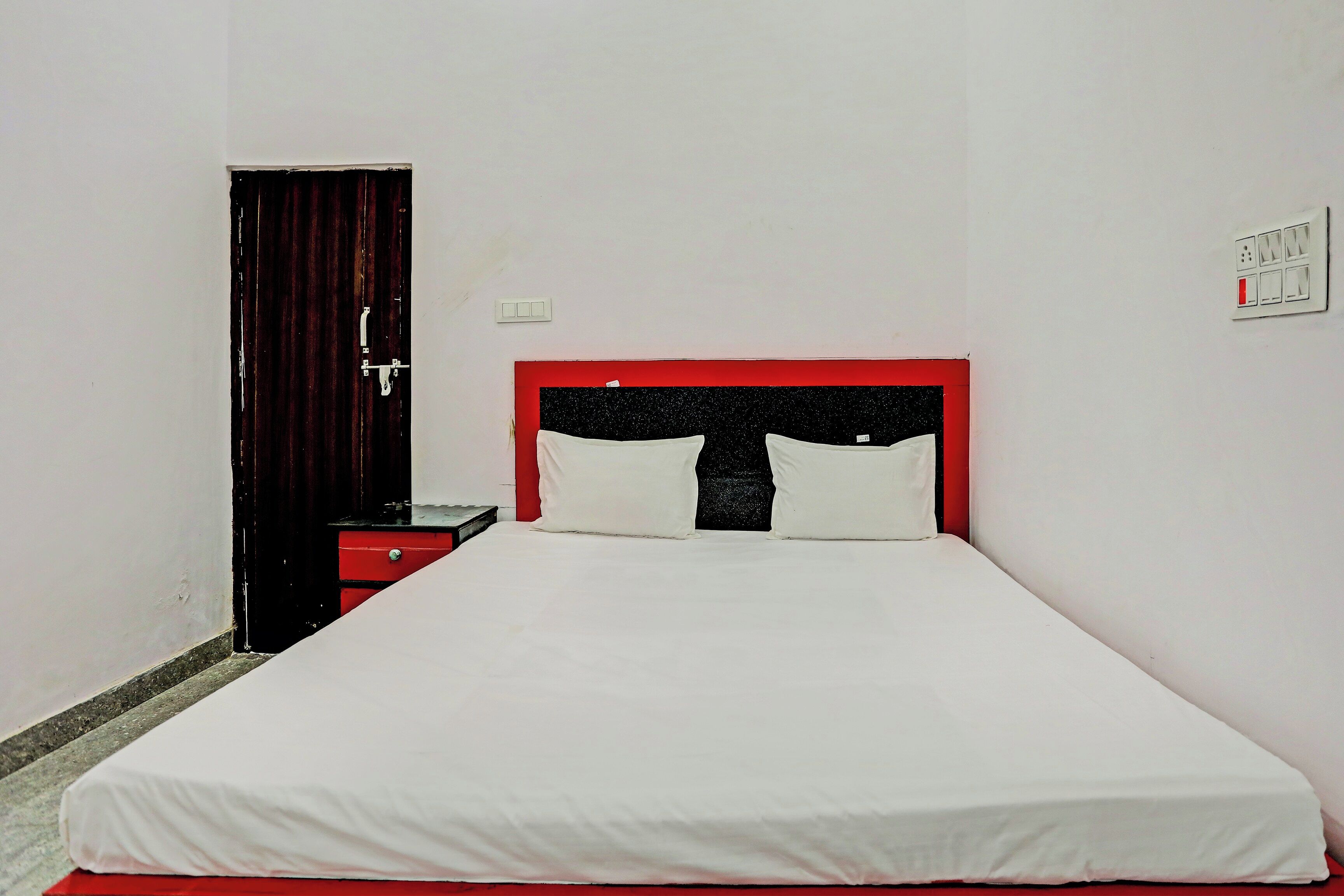 A single-bed room with a red headboard, white bedding, a nightstand, a red drawer, a door, and a wall-mounted light switch.