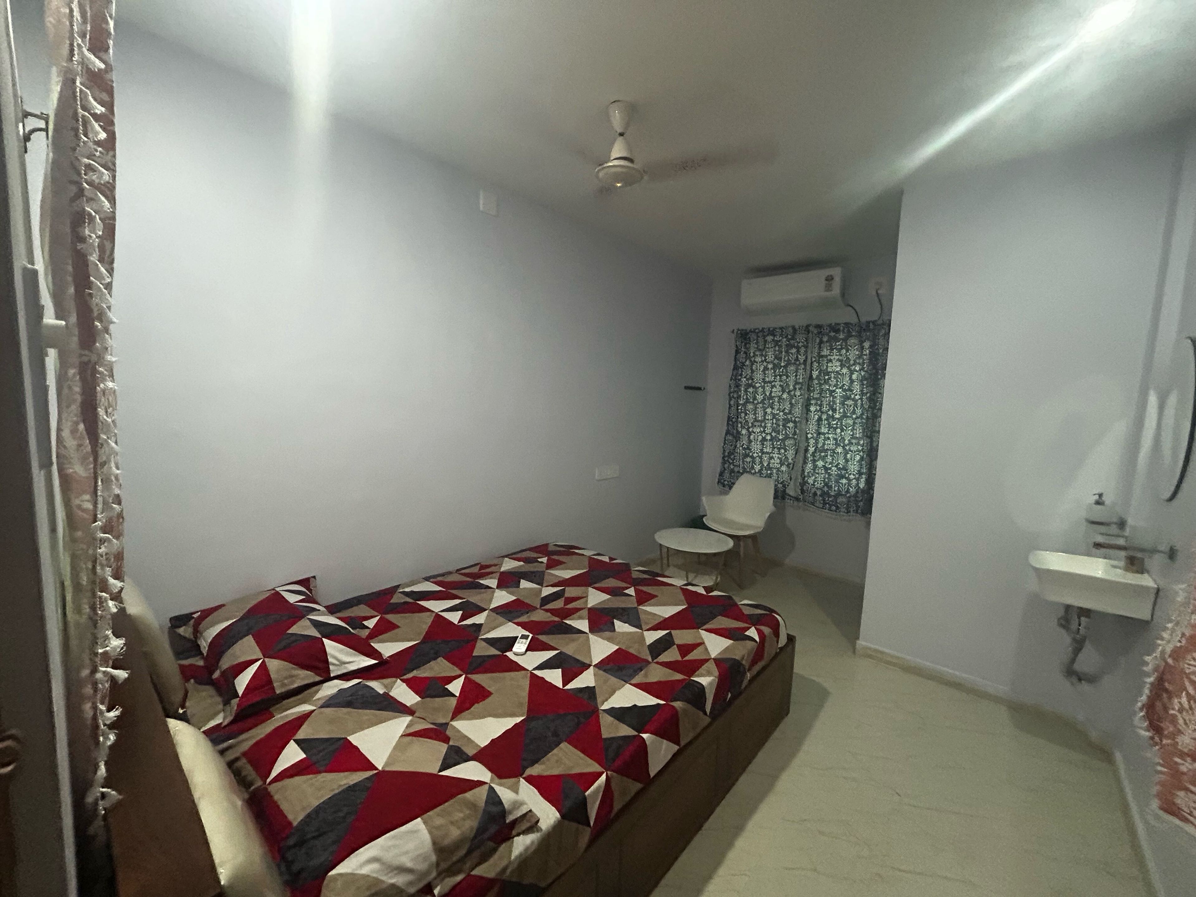 Una habitación pequeña con cama individual, colcha de diseño geométrico, ventilador de techo, una mesa pequeña con una silla y una ventana con cortinas estampadas.