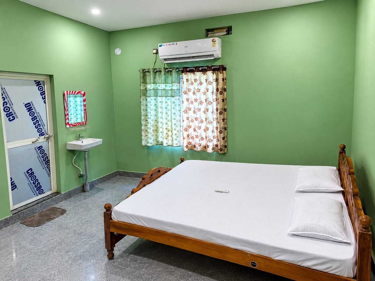 Un dormitorio con una cama, aire acondicionado de pared, una ventana con cortinas florales, un lavamanos y una puerta con espejo.
