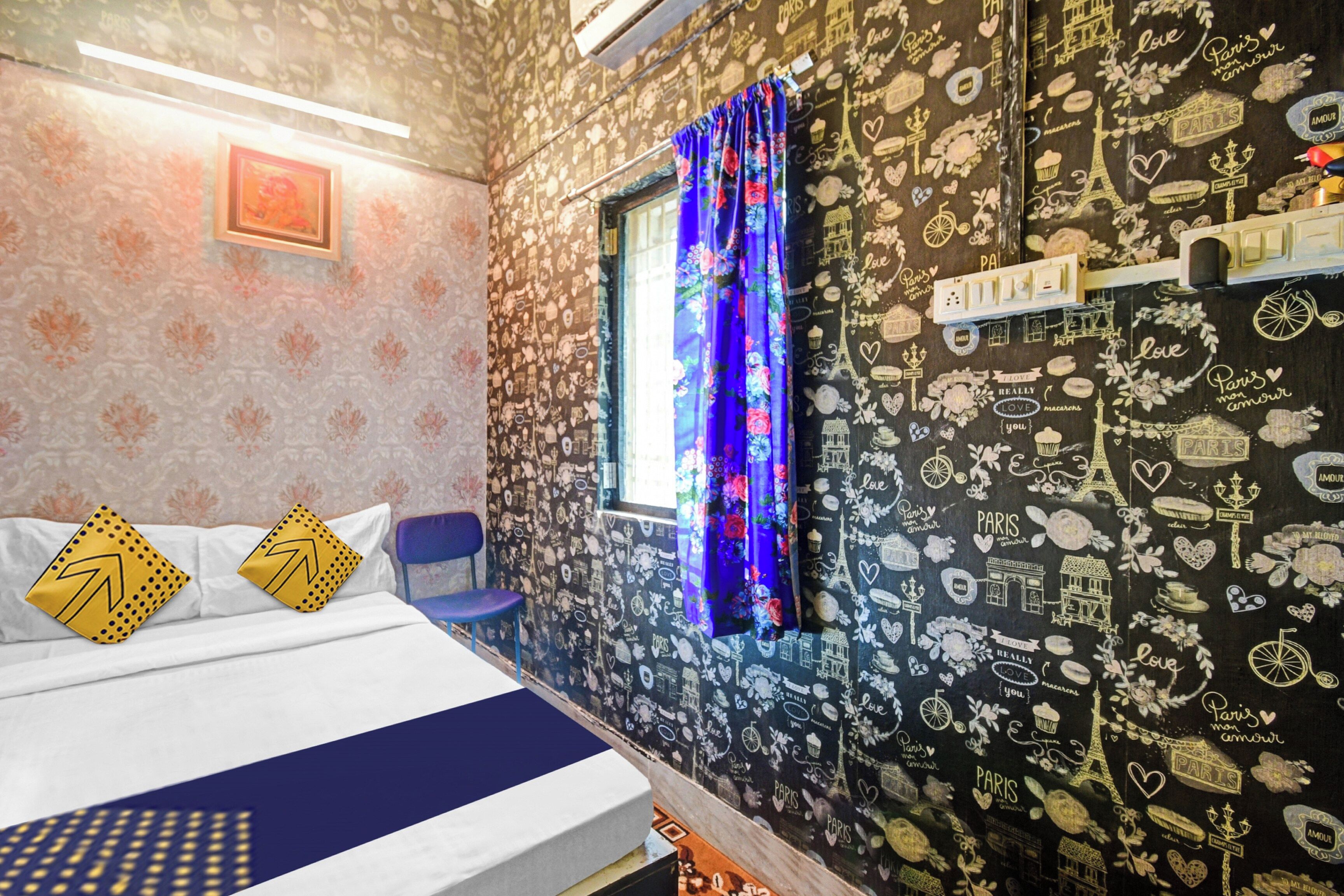 A hotel room with a bed, a window with curtains, a wall with Paris-themed decorations, and a small table with a lamp.