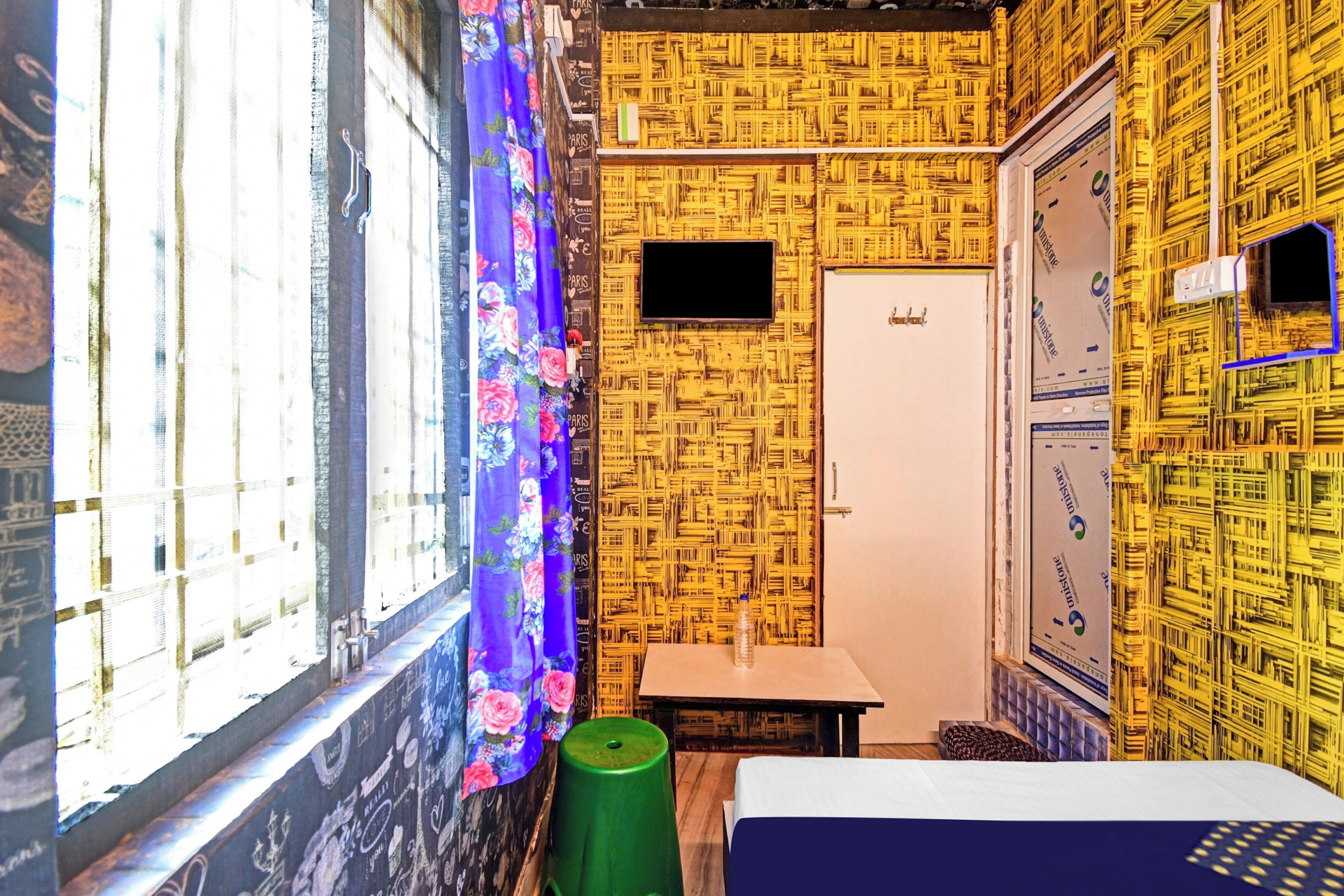 Quarto com papel de parede estampado, uma cama, uma mesinha, um banquinho verde e uma janela com cortina.