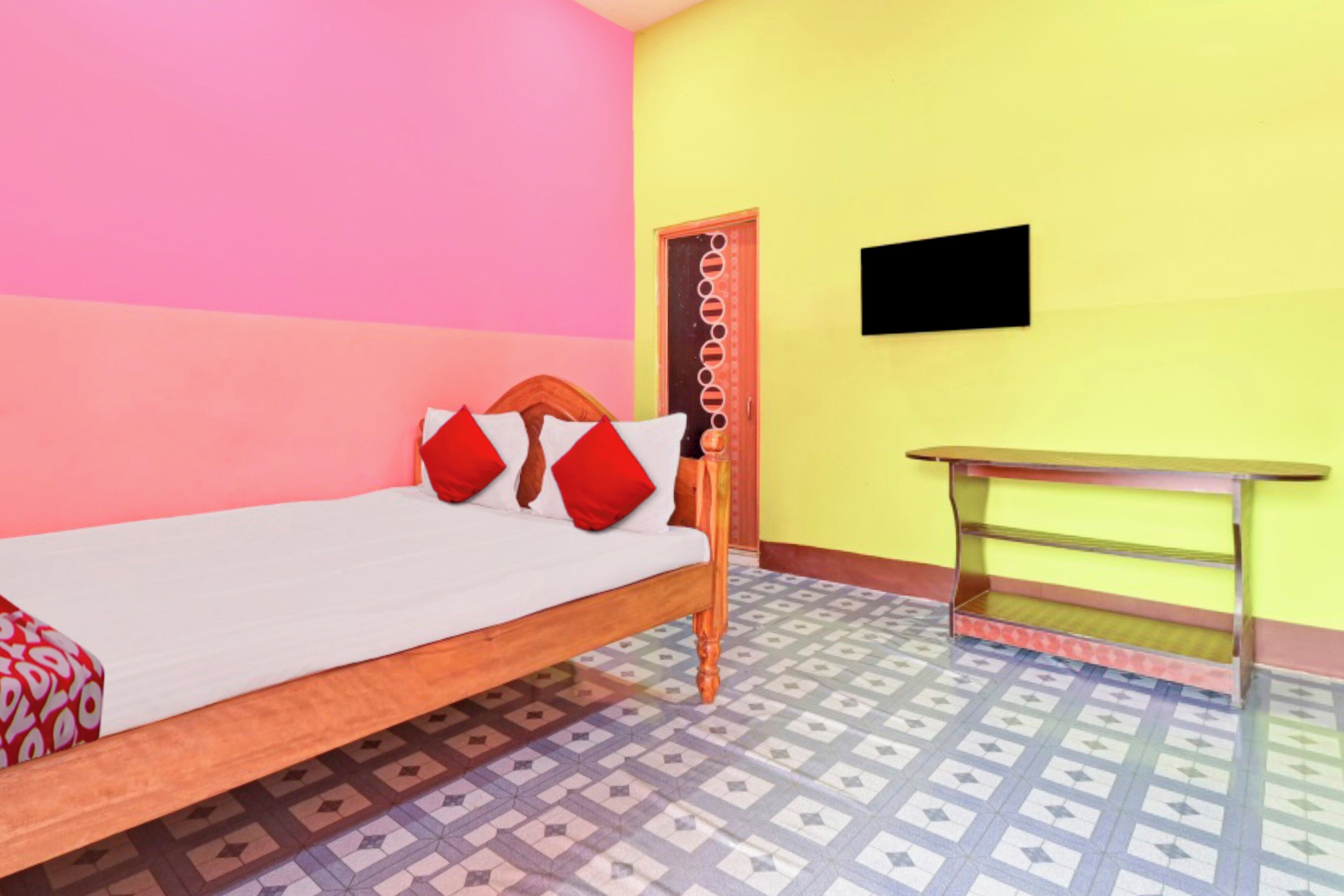 A single bed with a wooden frame and red pillows, a small table with a TV on the wall, and patterned floor tiles.