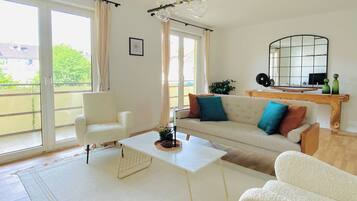 Ein modernes Wohnzimmer mit einer Couch, Sesseln, einem Couchtisch und einem großen Fenster mit Vorhängen.