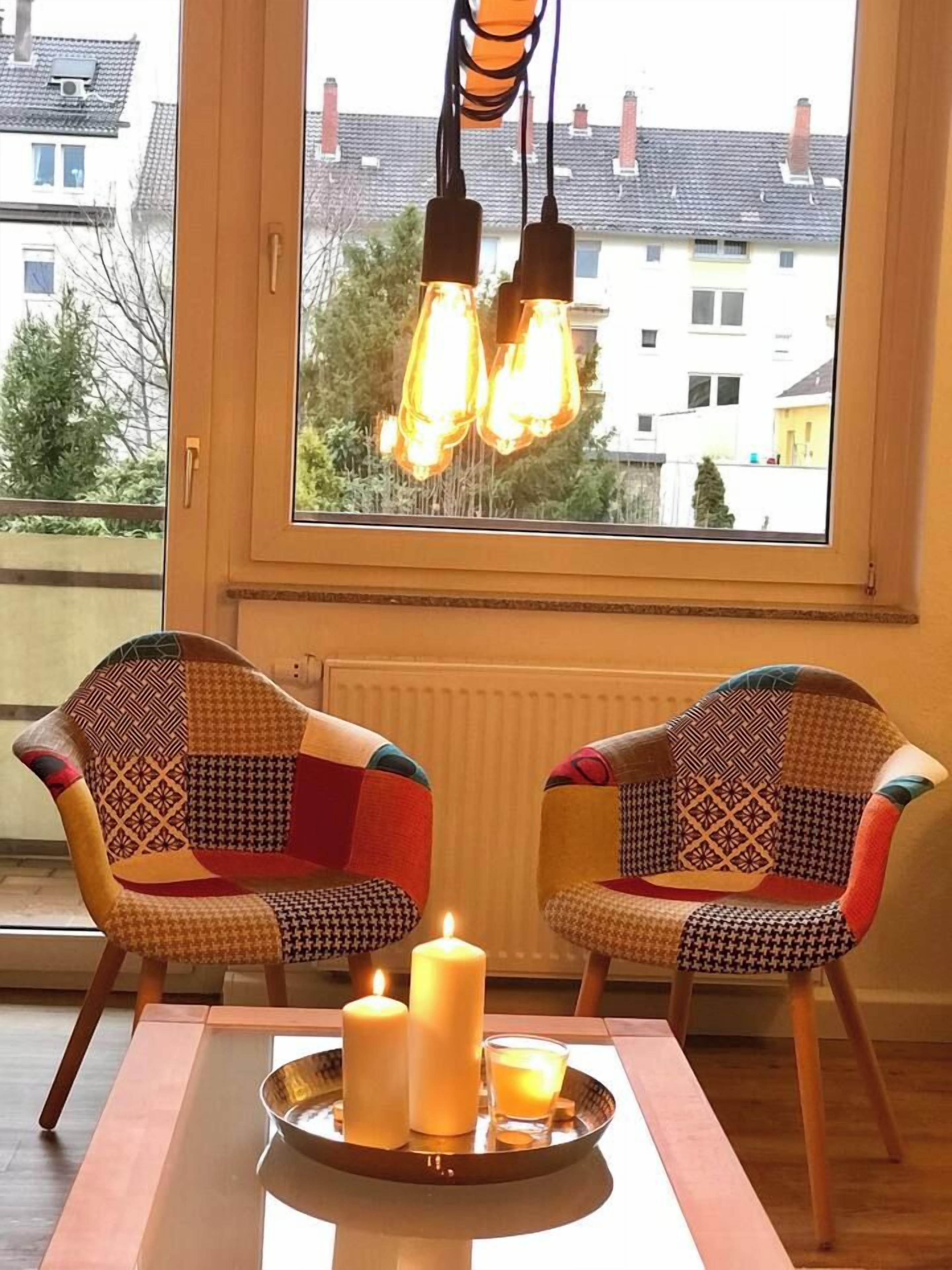 Zwei gemusterte Stühle, ein Tisch, Kerzen und ein Fenster mit hängenden Glühbirnen.