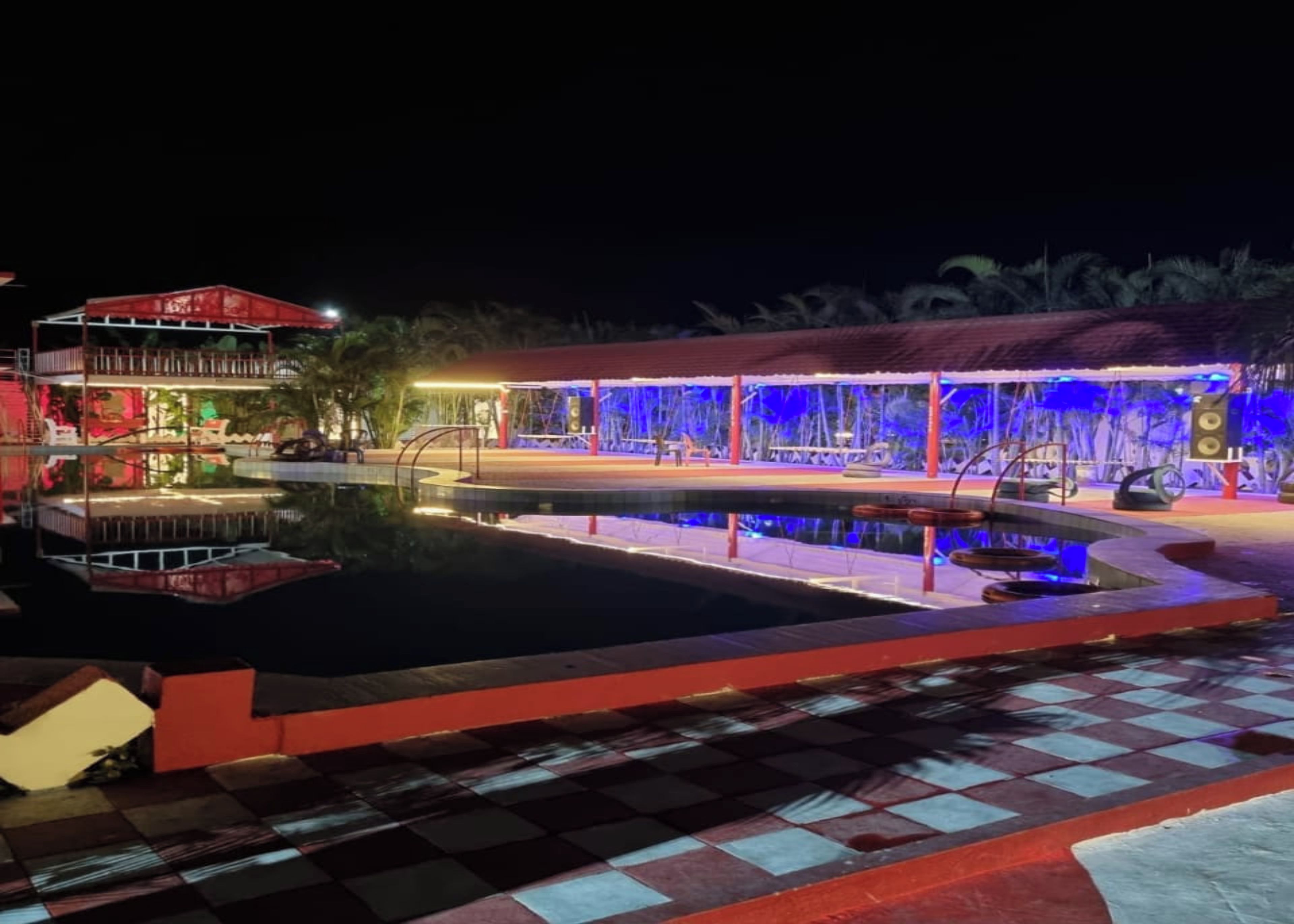 Vue nocturne d’un espace piscine avec un sol à carreaux, un coin salon abrité et un bâtiment à la façade rouge et blanche.