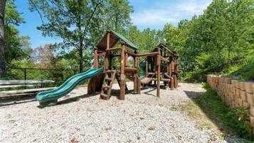 A playground with a slide and climbing structures, surrounded by trees and gravel.