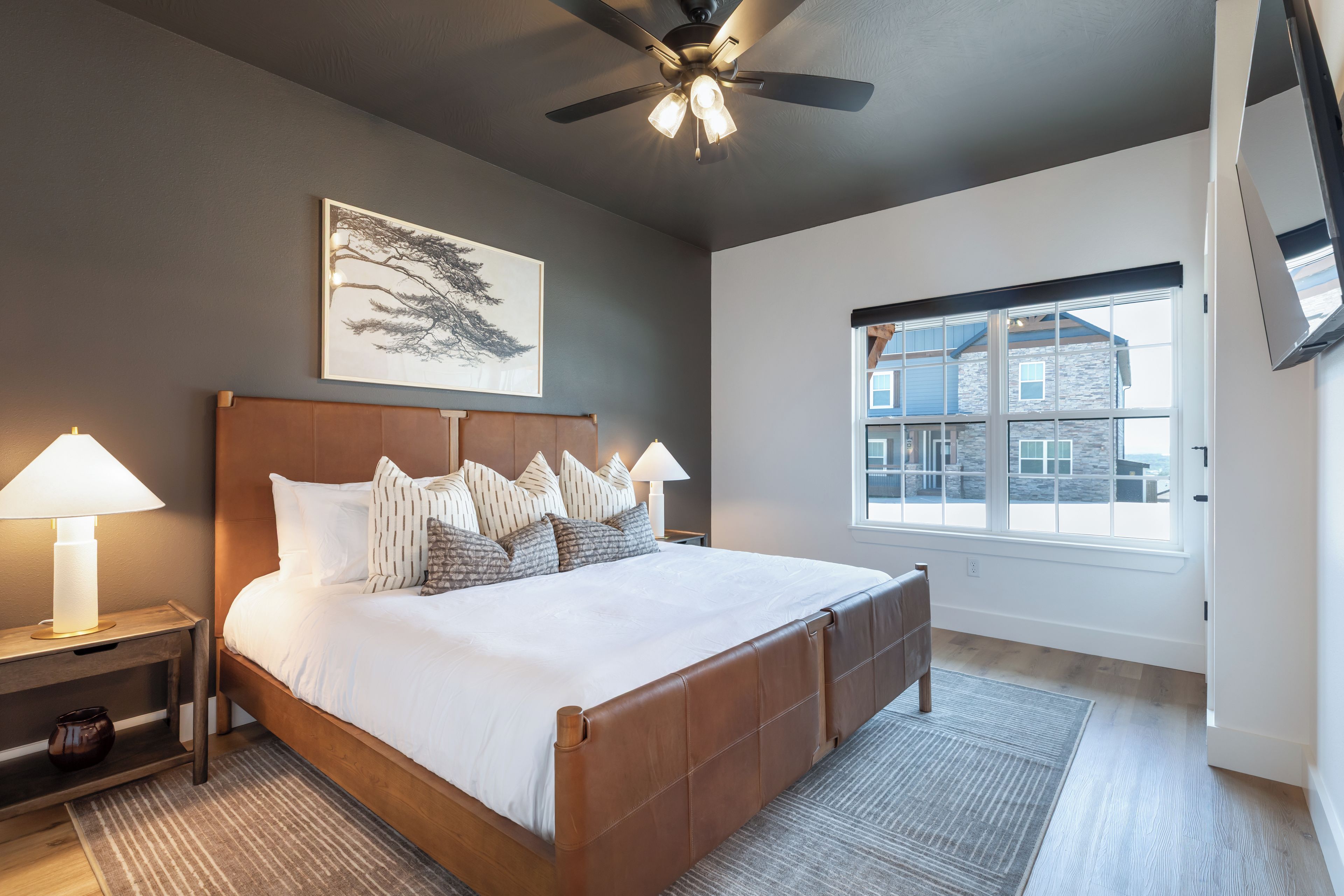 A bedroom with a large bed, a ceiling fan, a window with a view of buildings, and a wall-mounted artwork.