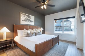 Un dormitorio con una cama grande, un ventilador de techo, una ventana con vistas a edificios y una obra de arte colgada en la pared.