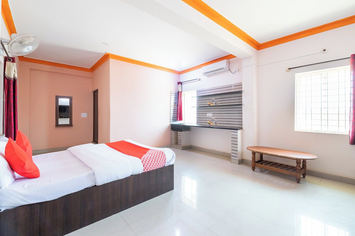 A hotel room with a bed, a bedside table, a window with curtains, an air conditioning unit, and a wall-mounted shelf.