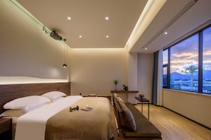 A modern hotel room with a large bed, a bench, a small table, and a view of a cityscape through a large window. - Ruyi Meiju Hotel (Lijiang Dashuiche) (Lijiang)