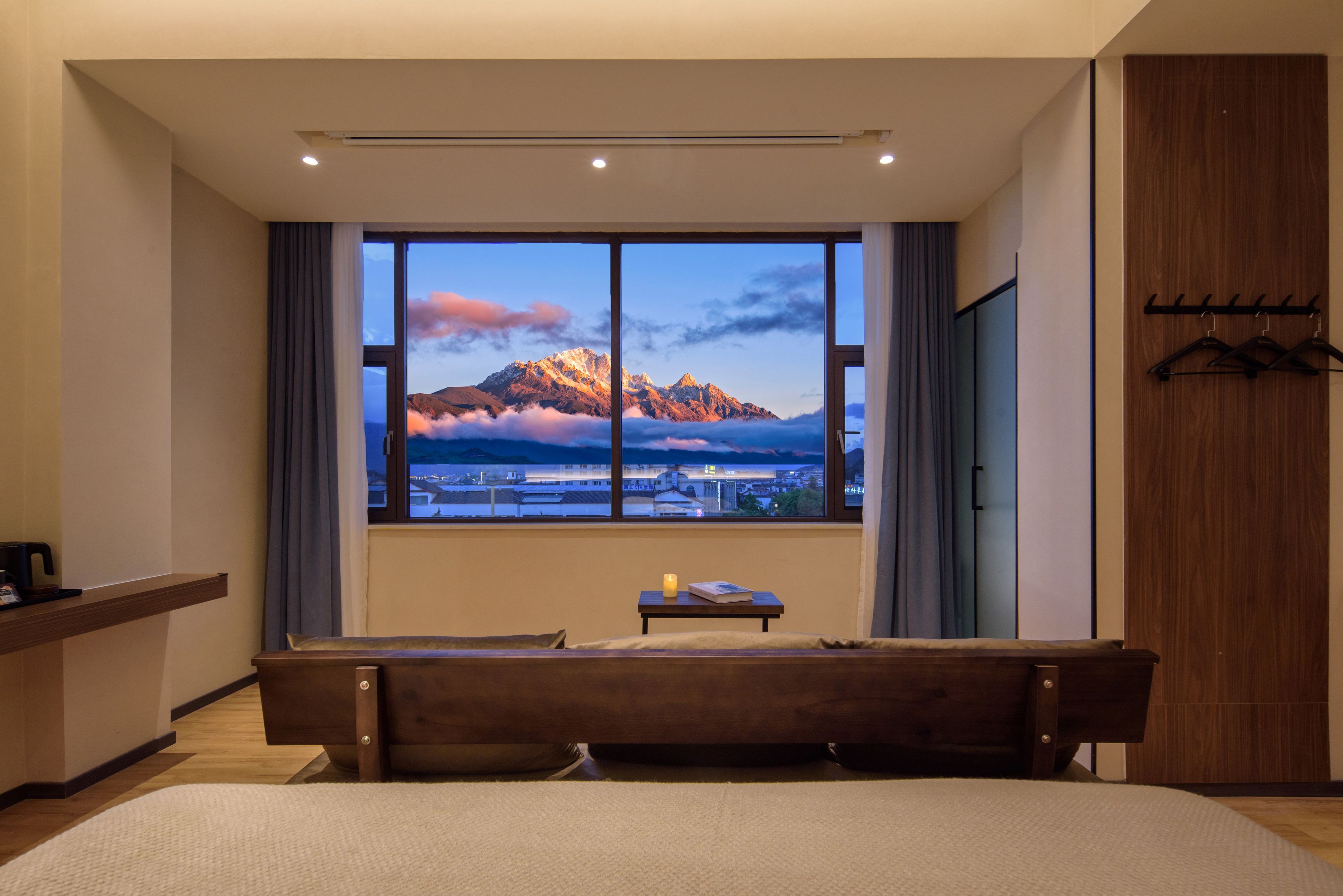 Habitación de hotel con un amplio ventanal que ofrece vistas a la montaña, un banco de madera, una pequeña mesa con un libro y un perchero.