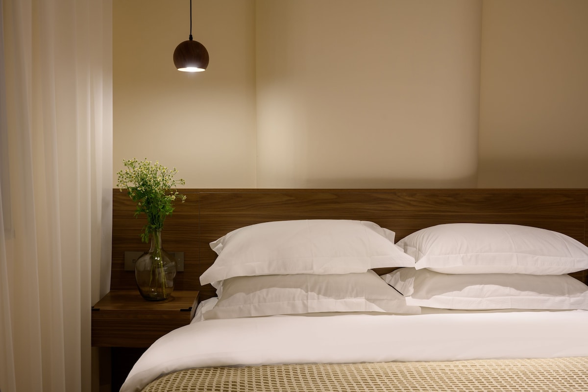 A neatly made bed with white linens, a wooden headboard, and a bedside table with a vase of flowers.