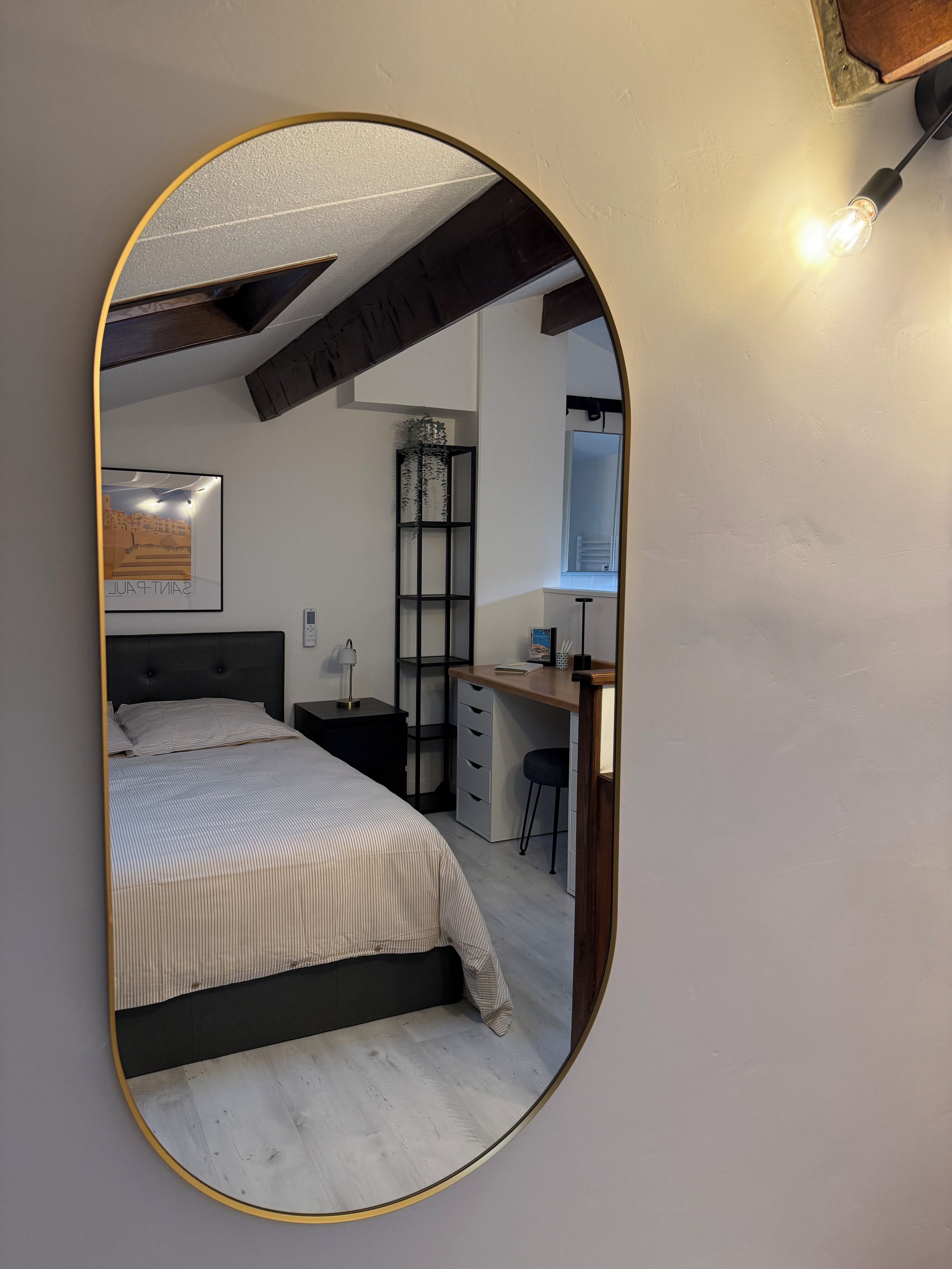 A bedroom with a bed, a desk, a chair, a nightstand, a mirror, and a wall-mounted light fixture.