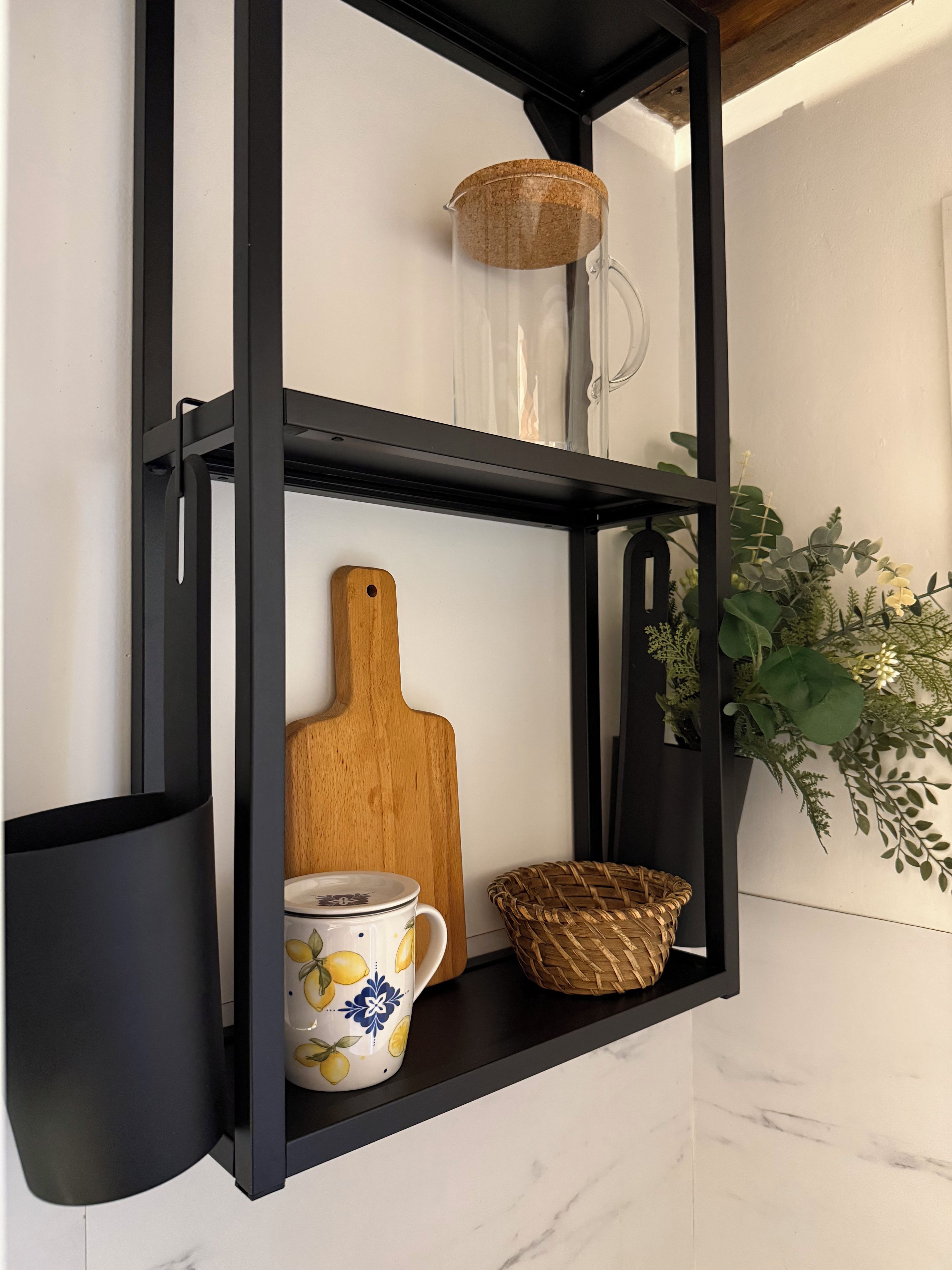 En svart metallhylla med en skärbräda i trä, en keramisk mugg och en flätad korg.
