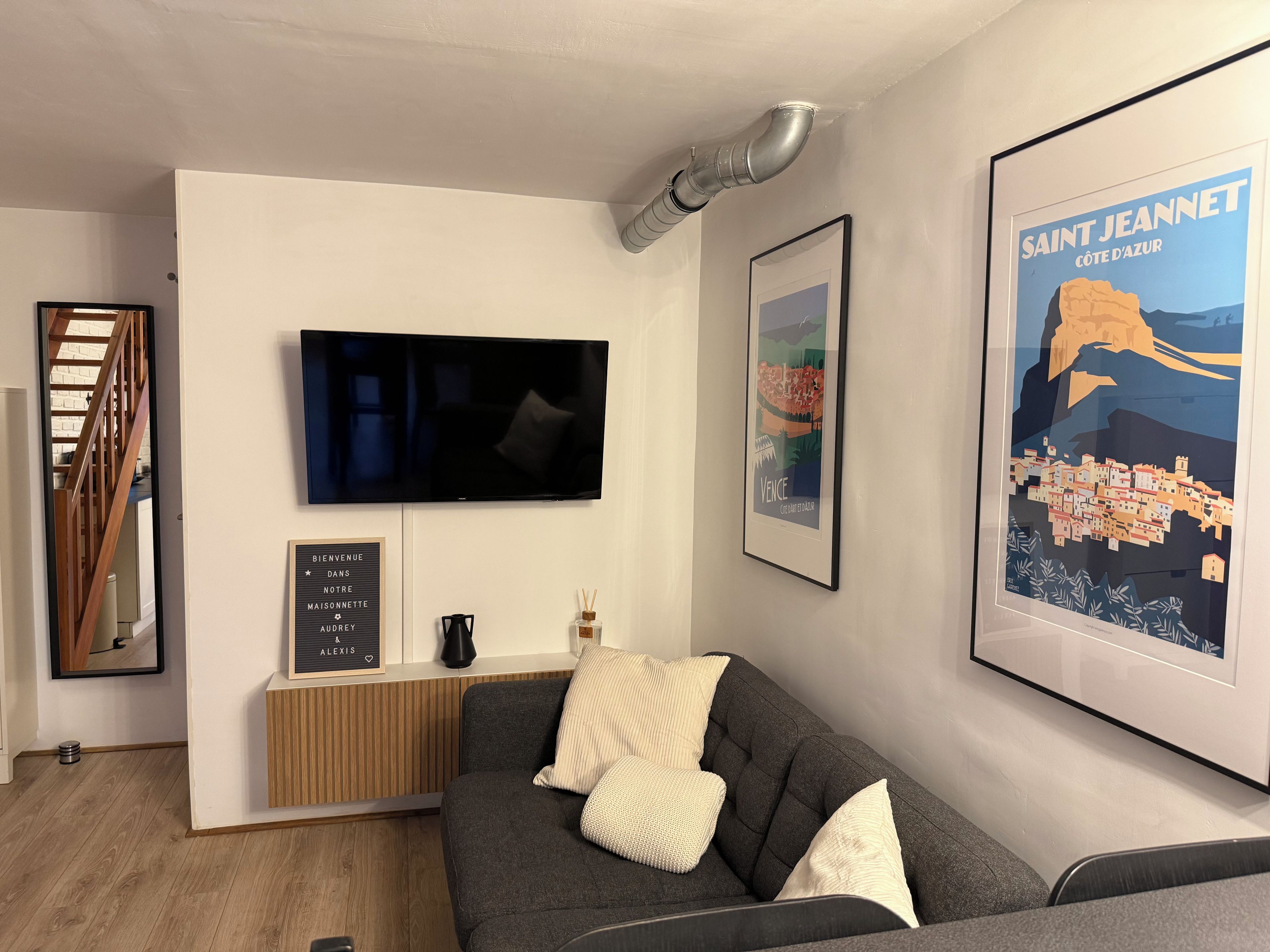 A modern living room with a flat-screen TV, a framed poster of Saint Jeannet, and a wooden bench.