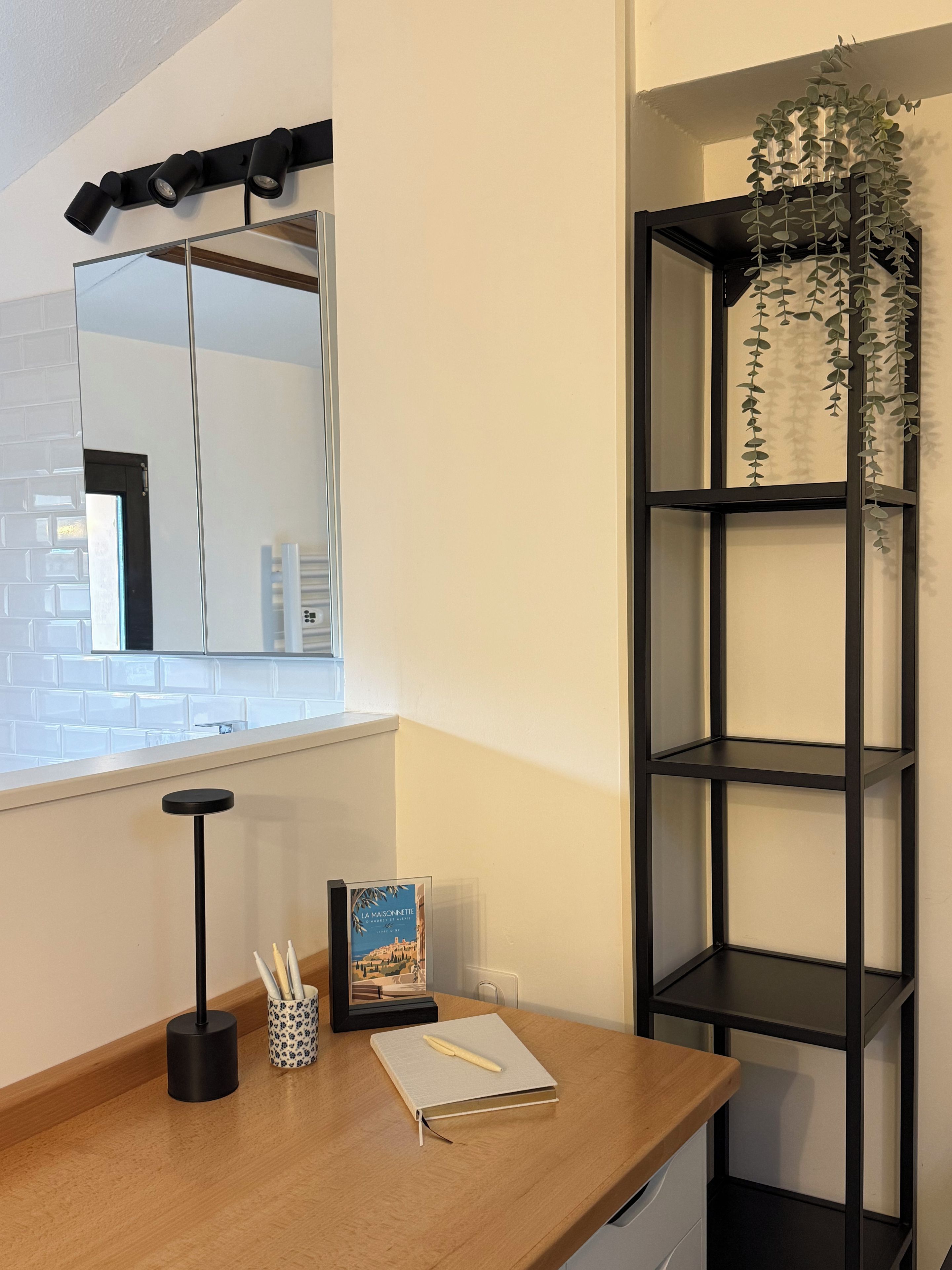 A wooden desk with a black lamp, a framed picture, and a notebook. A black shelf with a plant is to the right.