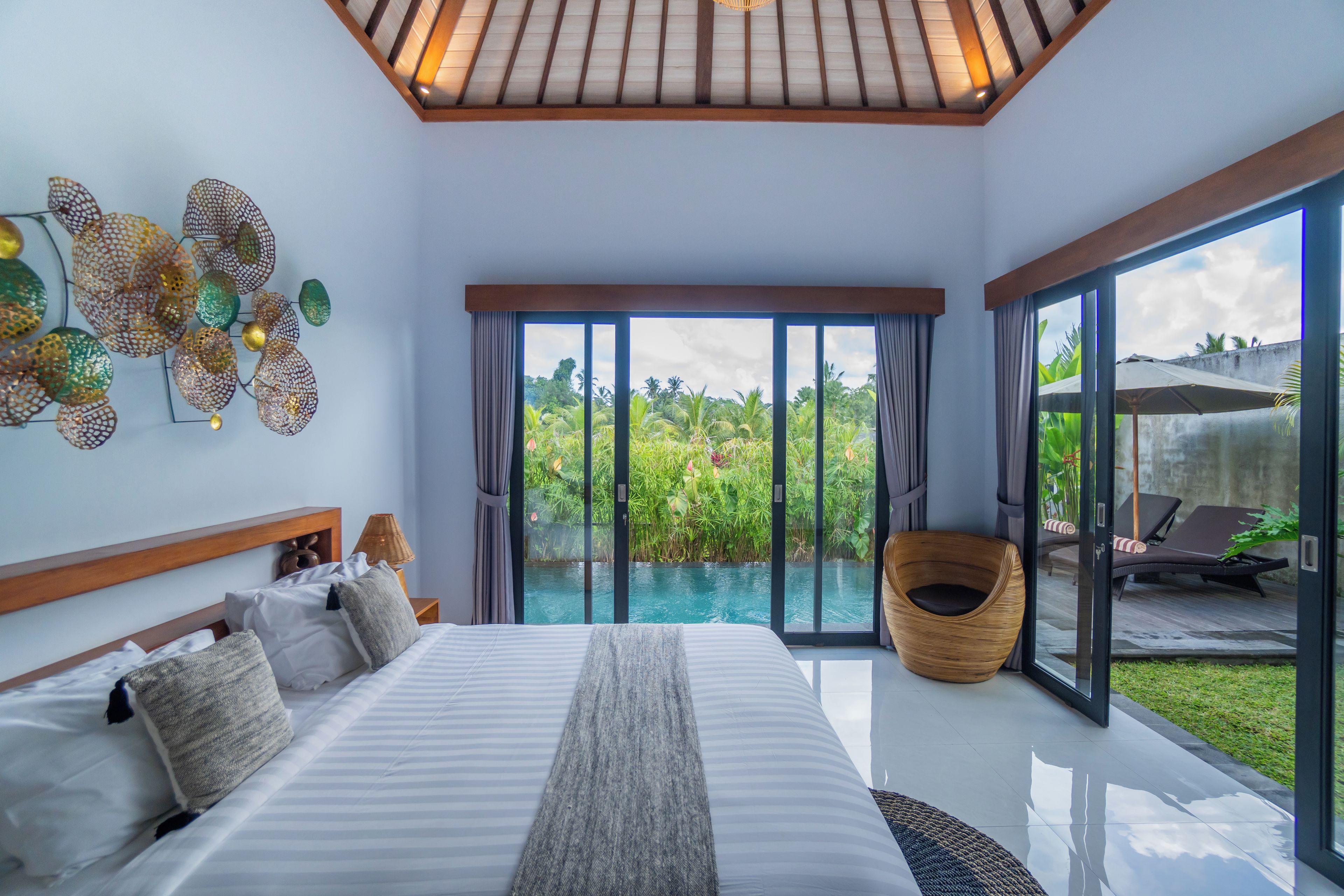 A modern bedroom with a large bed, a view of a swimming pool, and a balcony with lounge chairs.