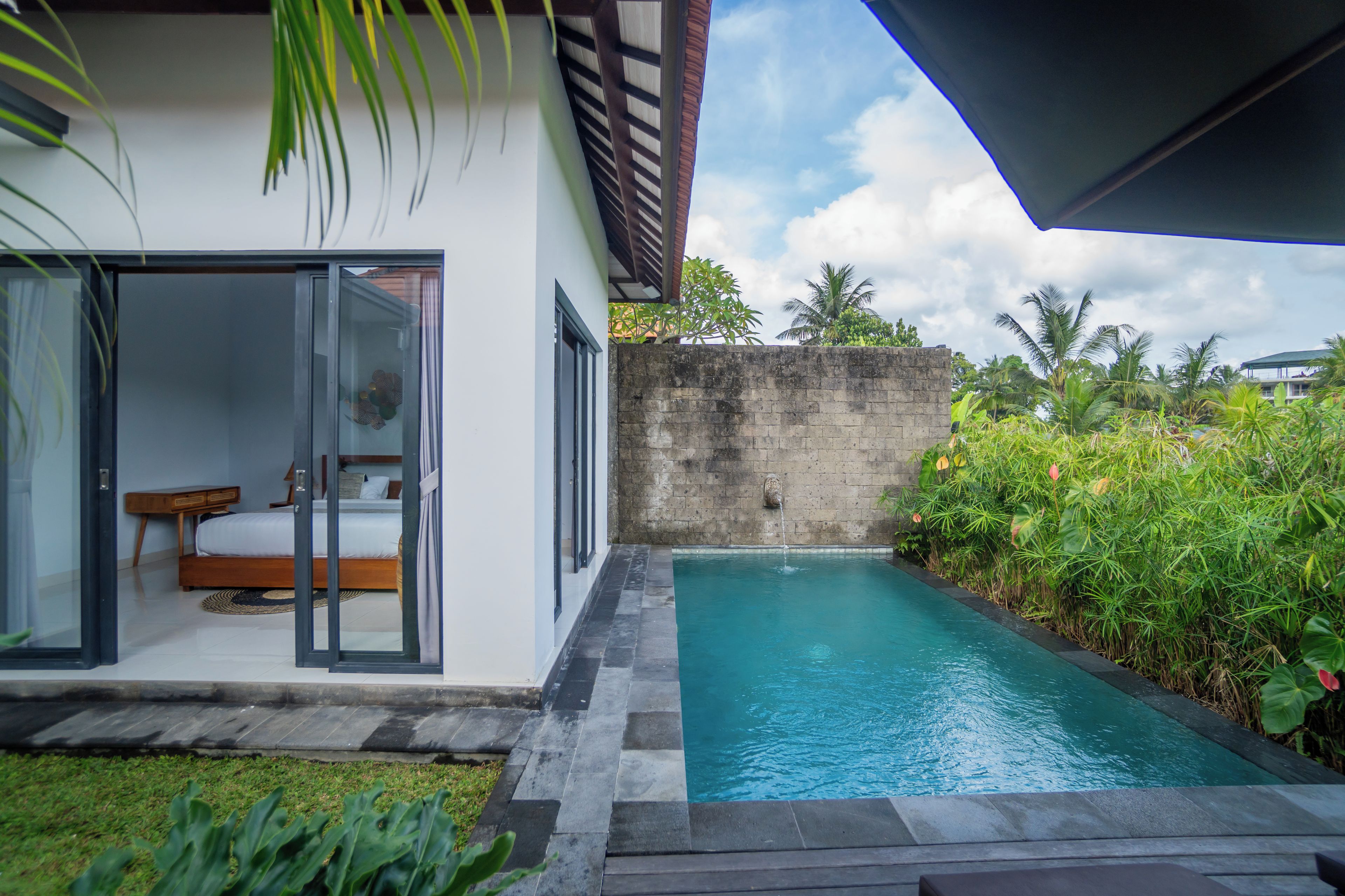Une maison moderne avec une porte vitrée donnant sur une chambre, une petite piscine et une végétation luxuriante.