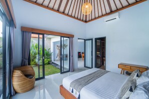Un dormitorio con una cama grande, cabecera de madera, una silla de mimbre y vistas a un jardín a través de puertas de cristal.