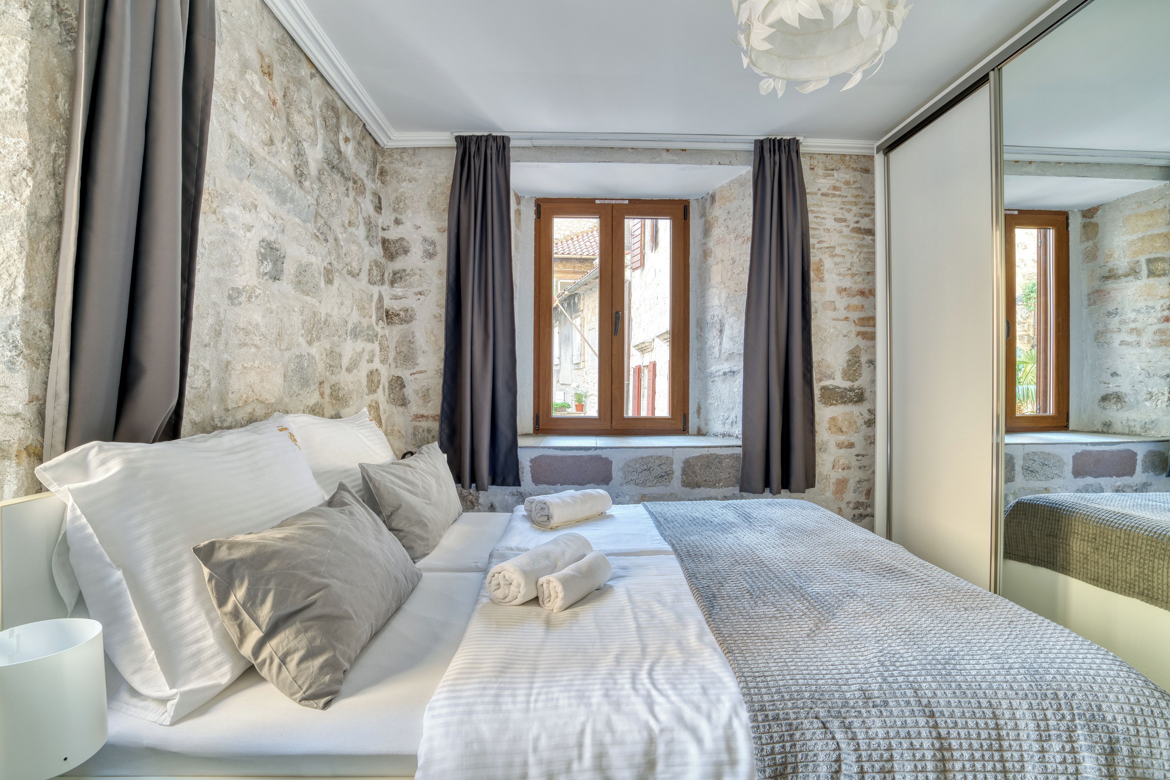 A bedroom with a stone wall, a bed with white bedding, a window with curtains, and a mirror reflecting the room.