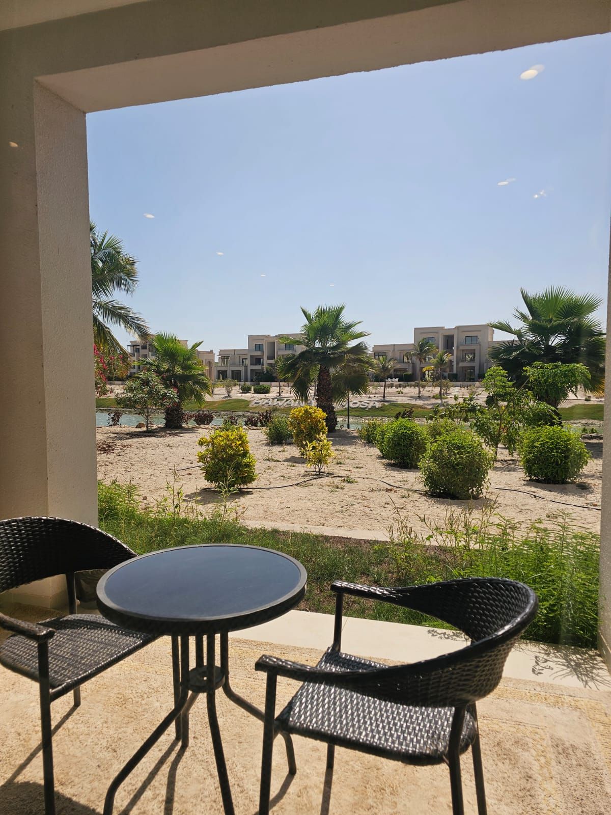 Un balcon agrémenté de deux fauteuils en osier et d’une table, donnant sur un espace paysager avec des palmiers et des arbustes, et un immeuble avec de nombreux balcons en arrière-plan.