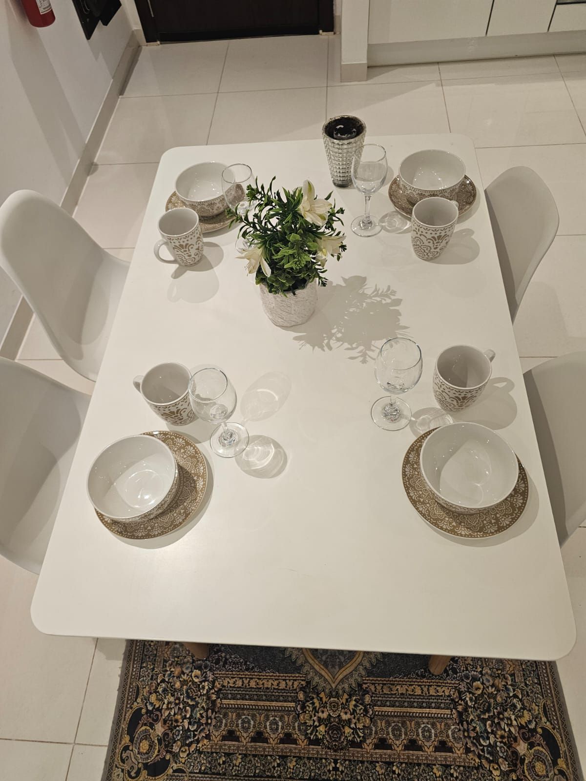 Una mesa de comedor dispuesta con platos, tazones y vasos blancos, rodeada de sillas blancas.