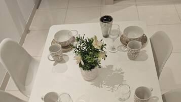 Una mesa de comedor dispuesta con platos, tazones y vasos blancos, rodeada de sillas blancas.