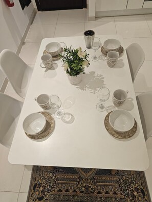 Una mesa de comedor dispuesta con platos, tazones y vasos blancos, rodeada de sillas blancas.