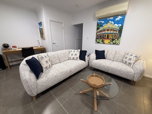 A living room with two sofas, a glass coffee table, a desk, and a painting on the wall. - Oasis Apartments Townsville (Garbutt)