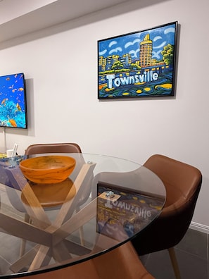 A dining area with a glass table, brown chairs, and a framed artwork on the wall. - Oasis Apartments Townsville (Garbutt)