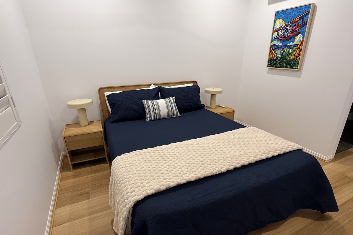 A neatly made bed with a dark blue bedspread and a cream-colored blanket, flanked by two bedside tables with lamps, in a room with wooden flooring and a single wall-mounted painting.