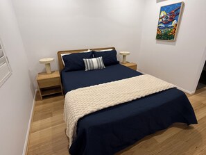 A neatly made bed with a dark blue bedspread and a cream-colored blanket, flanked by two bedside tables with lamps, in a room with wooden flooring and a single wall-mounted painting. - Oasis Apartments Townsville (Garbutt)